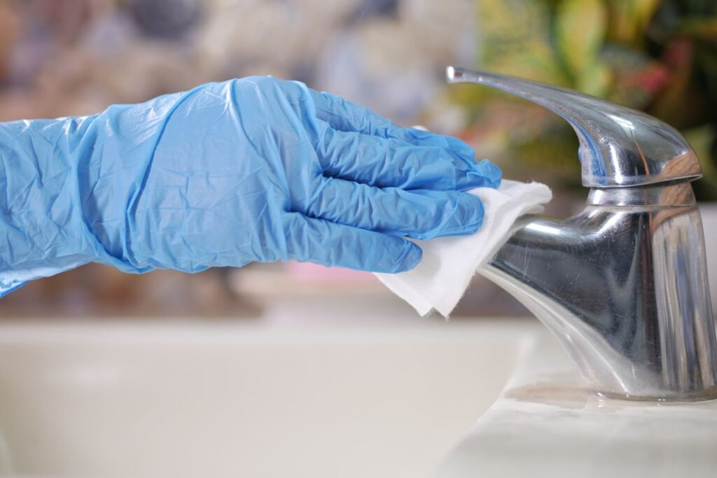 Close-up of gloved hand wiping a stainless steel faucet with a cloth indoors.