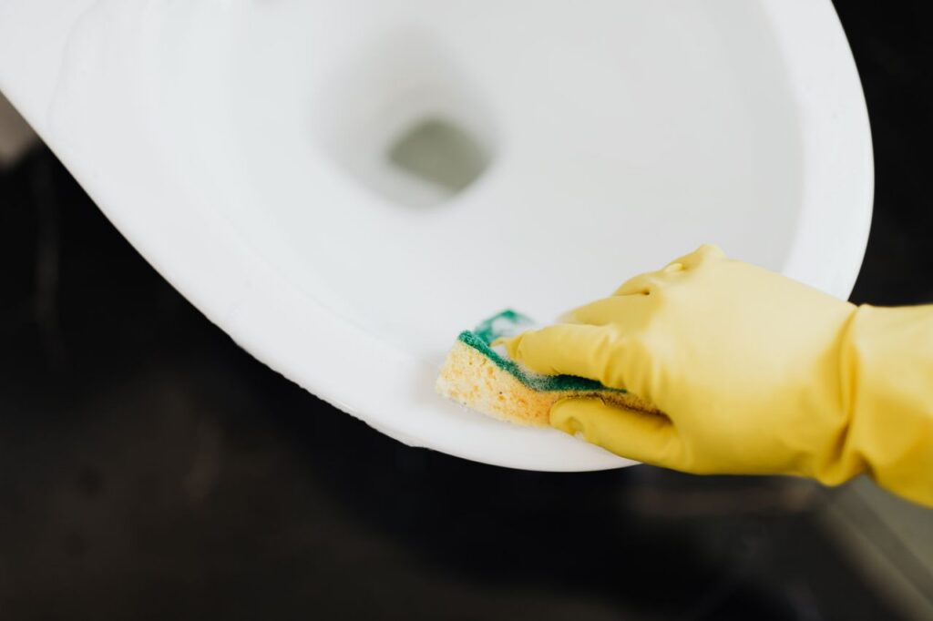 From above of crop unrecognizable person wearing yellow rubber gloves disinfecting and cleaning toilet bowl with sponge in bathroom
